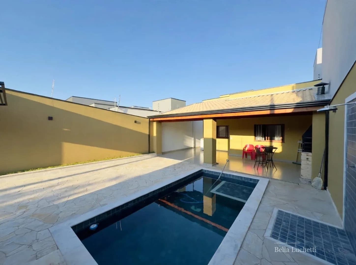 Quintal com piscina retangular de alvenaria, área coberta com churrasqueira e mesa com cadeiras.